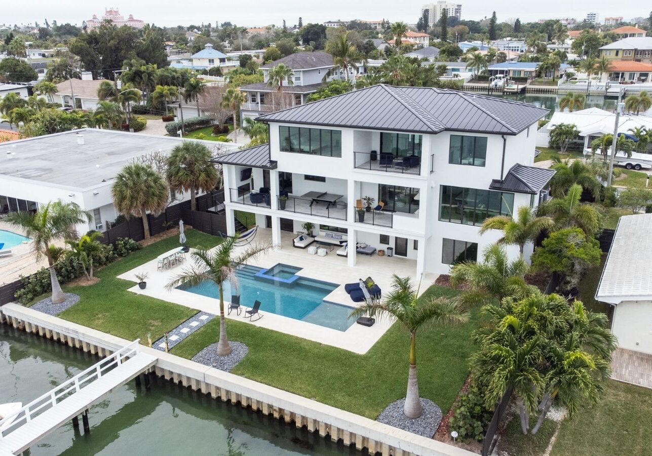 Aerial view of a modern waterfront luxury home featuring a swimming pool, landscaped garden, and outdoor seating area, surrounded by palm trees and a private dock on a serene canal.