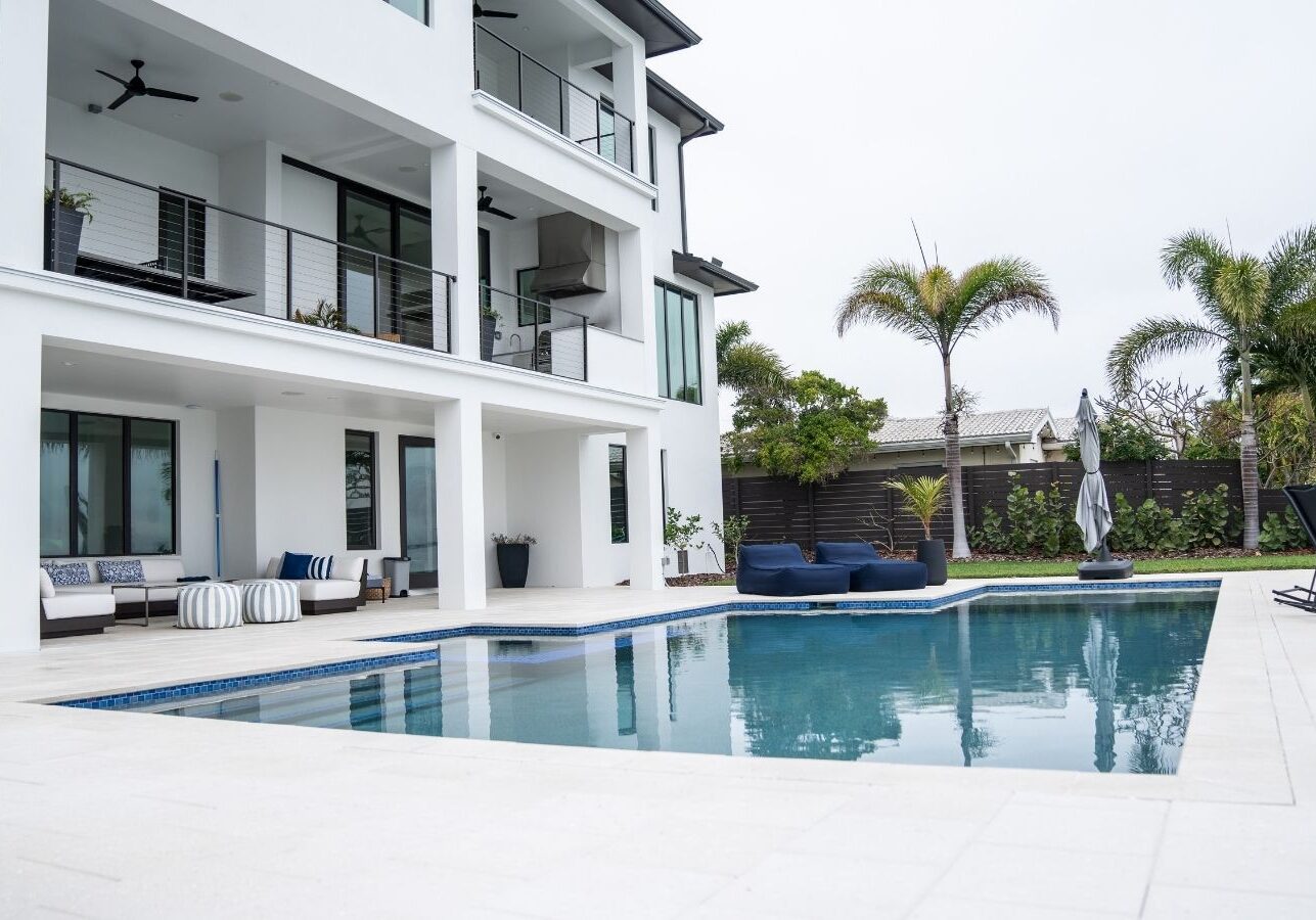 Modern luxury home with a swimming pool, featuring a spacious outdoor area, lounge furniture, and tropical landscaping. The two-story structure has large windows and a balcony, surrounded by palm trees, creating a serene atmosphere.