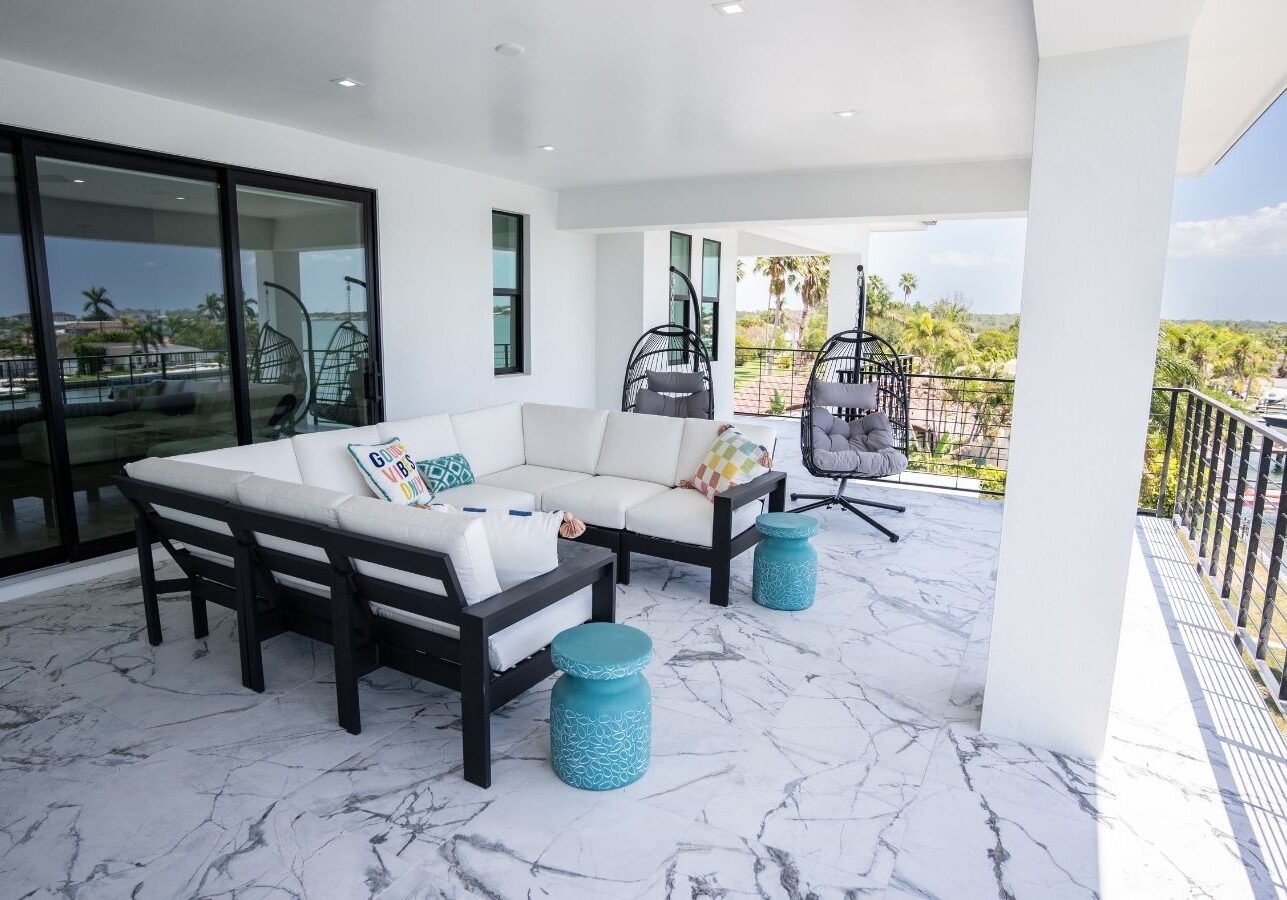 Modern outdoor patio featuring a stylish sectional sofa with decorative pillows, two hanging egg chairs, and turquoise accent tables, set against a backdrop of palm trees and a marina view. The space is illuminated by natural light, showcasing the elegant marble flooring and contemporary design.